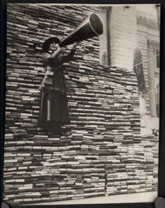 Image of librarian with megaphone in front of a wall of books donated for soldiers in WWI. 