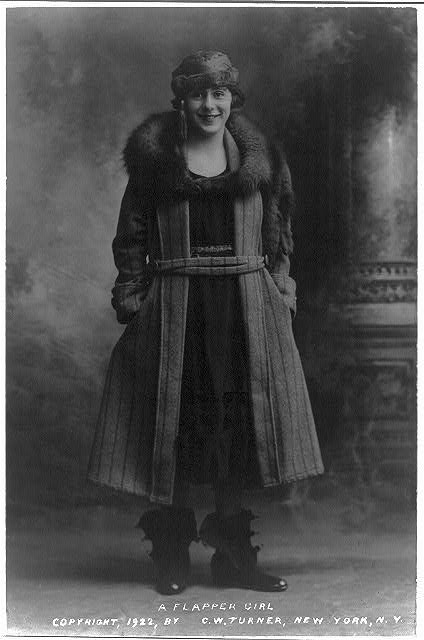 Photo of a young flapper dressed in a wool coat with a fur collar, a cloche hat with a tassel, and rubber galoshes with the clamps left open.