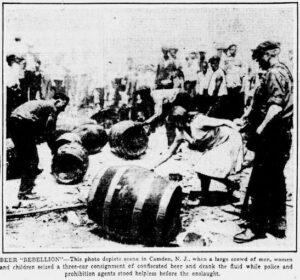 Black and white photograph of approximately 15 people and 5 scattered beer barrels, some of the people are reaching for the barrels to drink the beer, others are law enforcement looking on. The caption reads, "Beer Rebellion" and explains that the beer had been seized by a large crowd of men, women, and children, and that police and prohibition agents quote, "stood helpless before the onslaught".